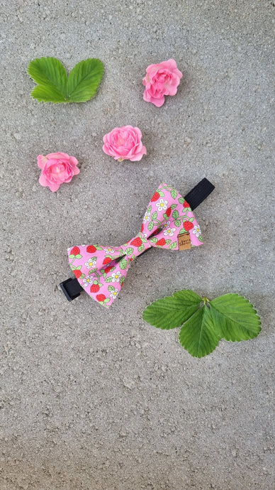 A dog bow tie with pink foundation, red strawberries, green leavea and white flowers.