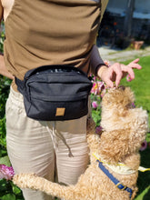 Load image into Gallery viewer, A person wearing a black fanny pack with an outside pocket where the zipper is hidden. A brown faux suede tag with the name Beans &amp; Sploot in the middle of the bag. The person is giving a treat from the treat bag to a dog who is standing with his paws on the person&#39;s legs.
